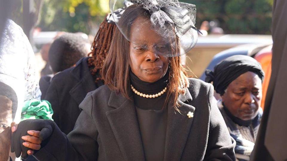 Esther Lungu, the former president's widow, at a memorial for her husband. Her eyes are cast down. She is wearing a black hat and black outfit and coat with a white beats.